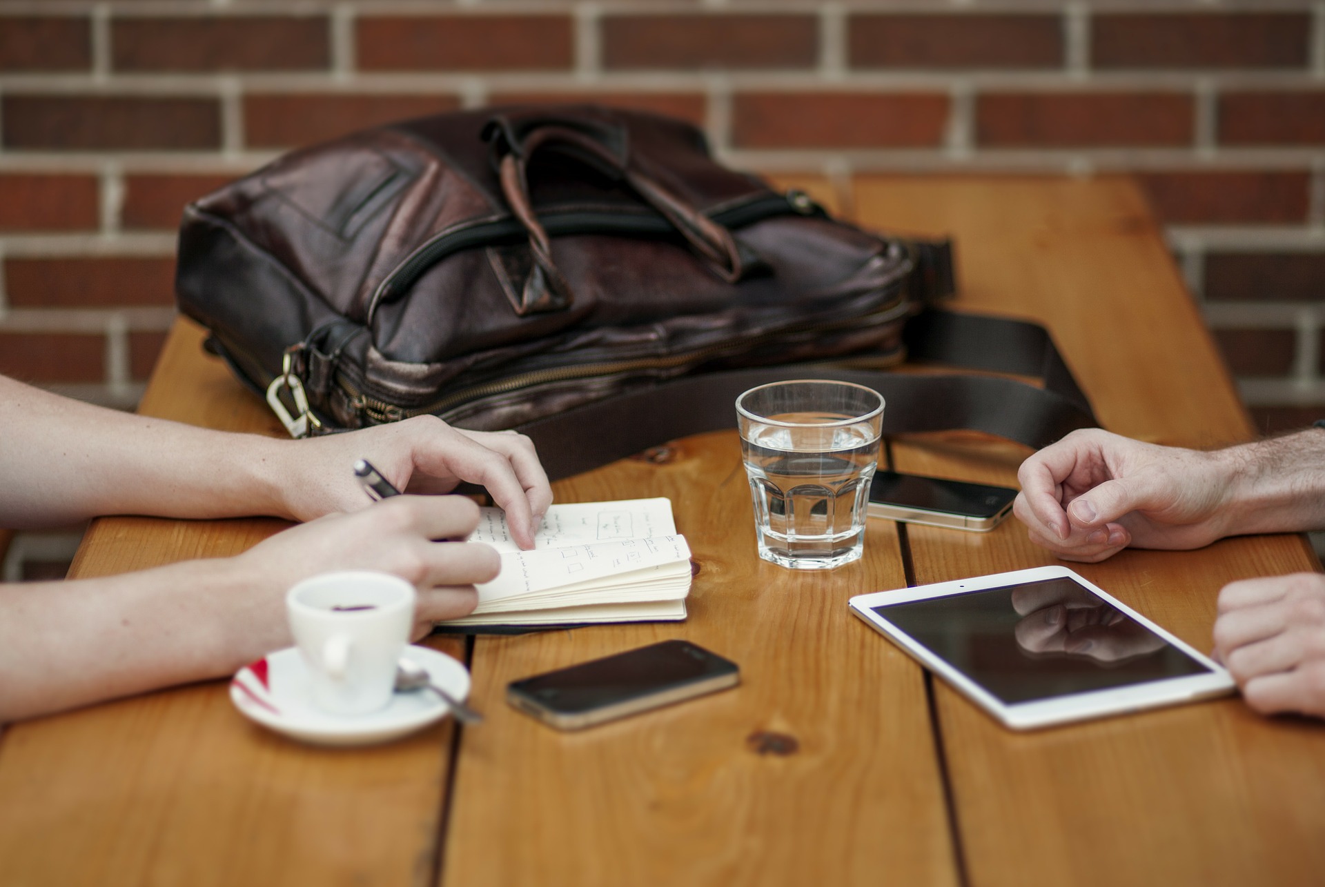 Zwei Personen sitzen an Holztisch mit Notizbuch, Tablet, Kaffee und Glas Wasser.